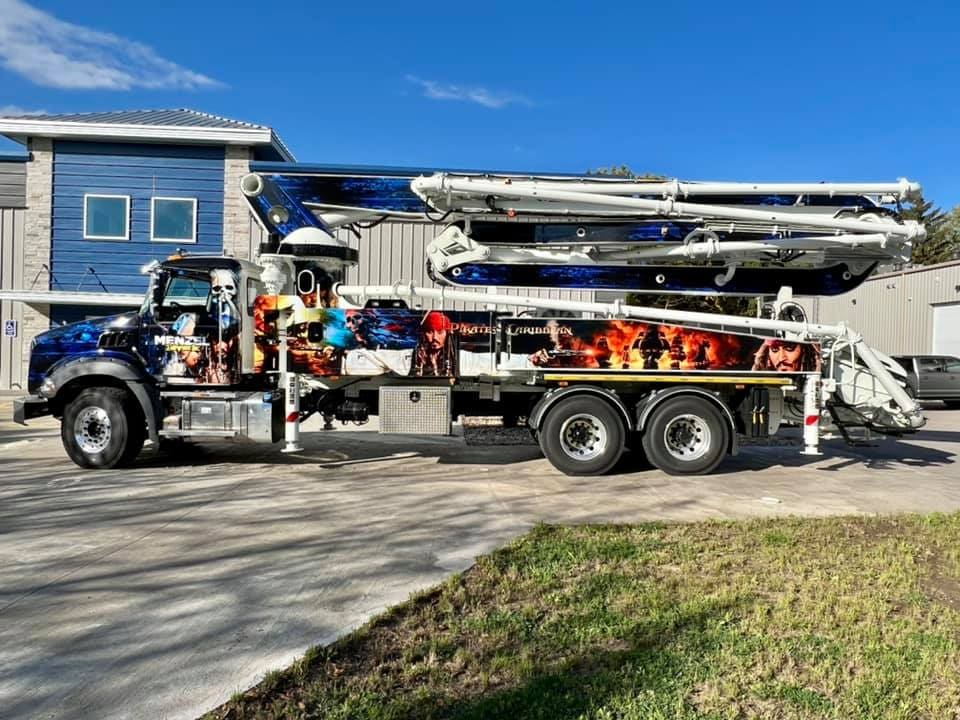 A large silver and blue tow truck featuring Menzel Towing Service LLC with a Pirate of the Caribbean and Jack Sparrow print on the side, it is parked on a concrete driveway