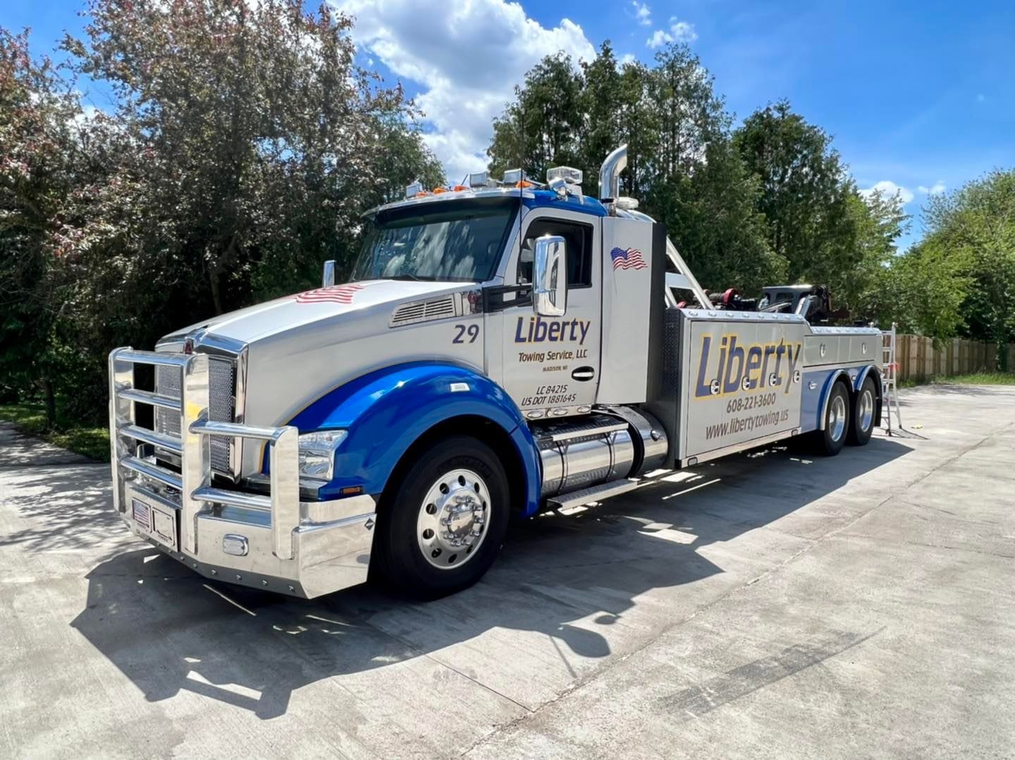 A large silver and blue tow truck featuring Truck Fleet Graphics Madison and Liberty Towing Service LLC on the side is parked on a concrete driveway, set against trees and a blue sky with scattered clouds.