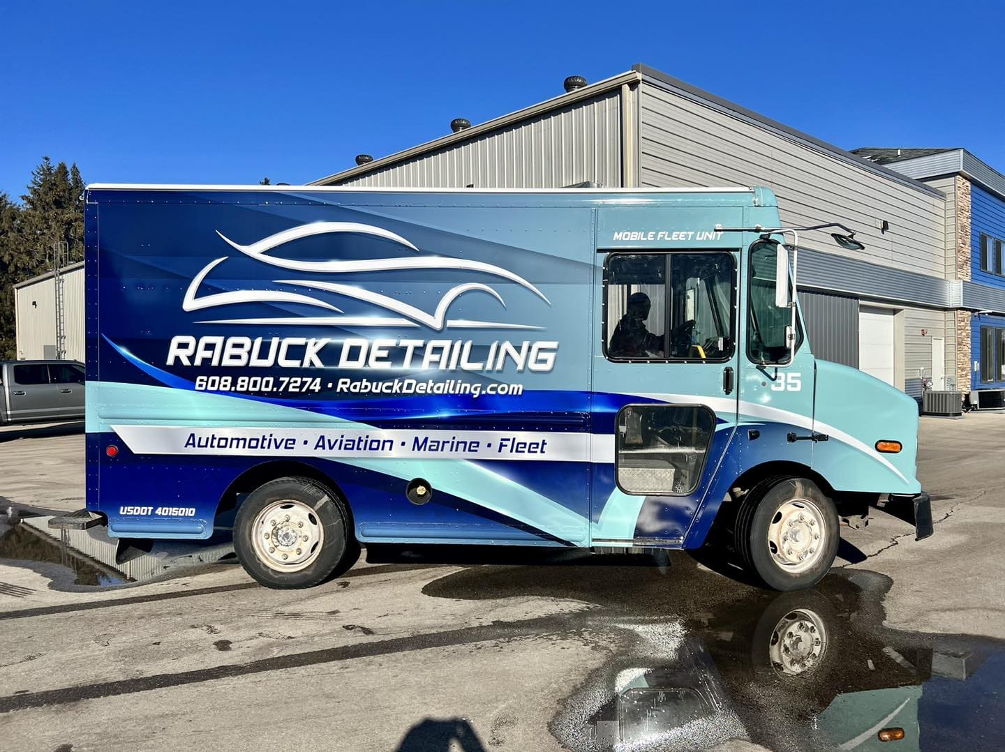 A box truck with RABUCK DETAILING and contact info in large white letters, parked outside a gray building. The blue and teal truck advertises automotive, aviation, marine, fleet detailing services, and eye-catching Fleet Wraps and Graphics.