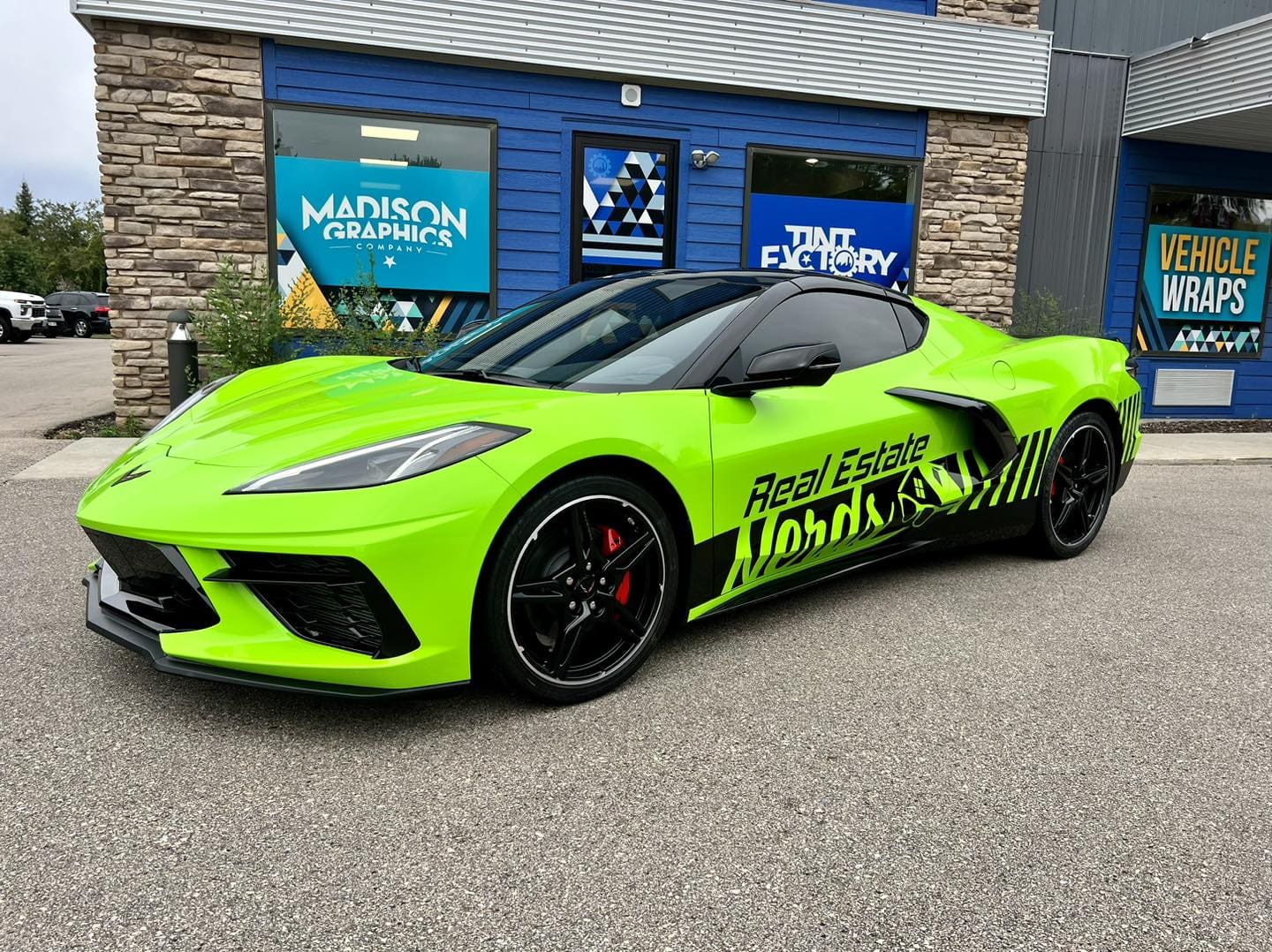 A bright green sports car with black rims and Real Estate lettering—showcasing creative vehicle wraps—is parked in front of the Madison Graphics Company building with stone and blue siding, featuring signs for graphics and tinting services.