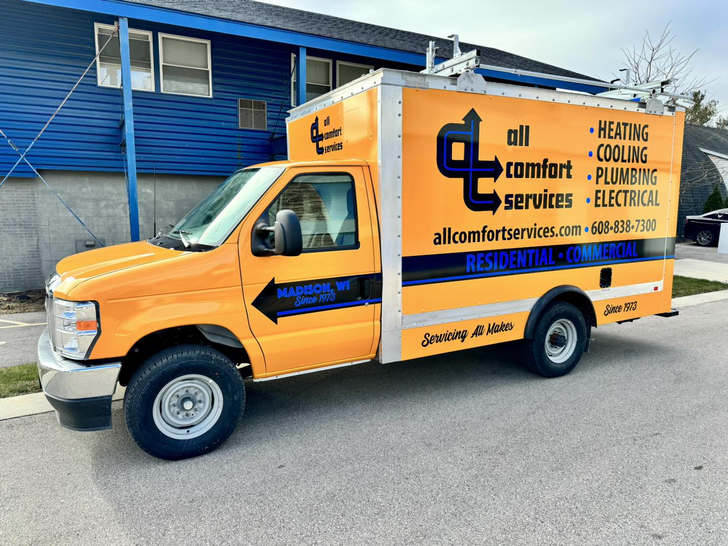 A yellow and blue All comfort services brothers hauling truck, promoting a HVAC and plumbing business with fleet graphics, is parked outside a building. The vehicle wrap features company details