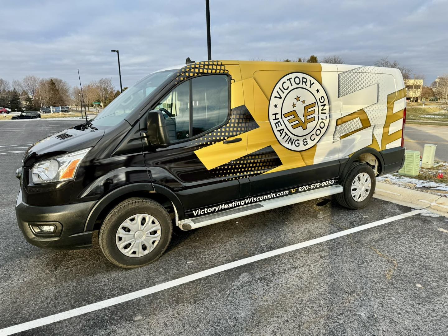 A black, gold, and white Victory Heating & Cooling van featuring custom fleet wraps and graphics is parked in a lot. The logo and website are visible on the side, with trees and buildings in the background.