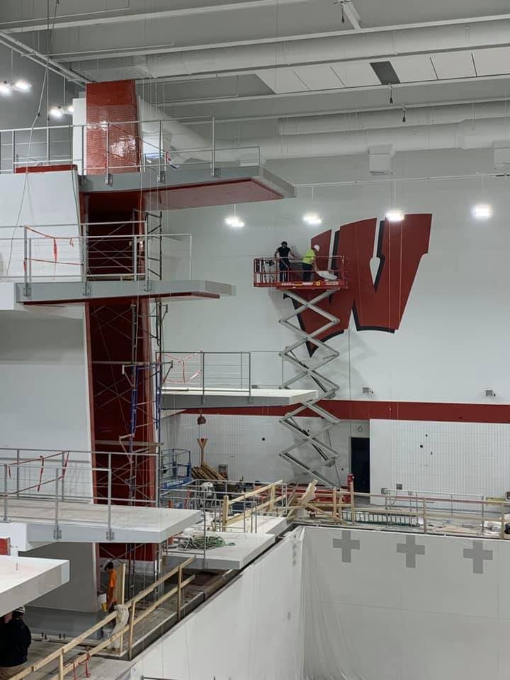 Workers on a scissor lift putting up a wall mural of the UW-Madison W Logo in the Aquatic Center while construction is being performed without water in the pool
