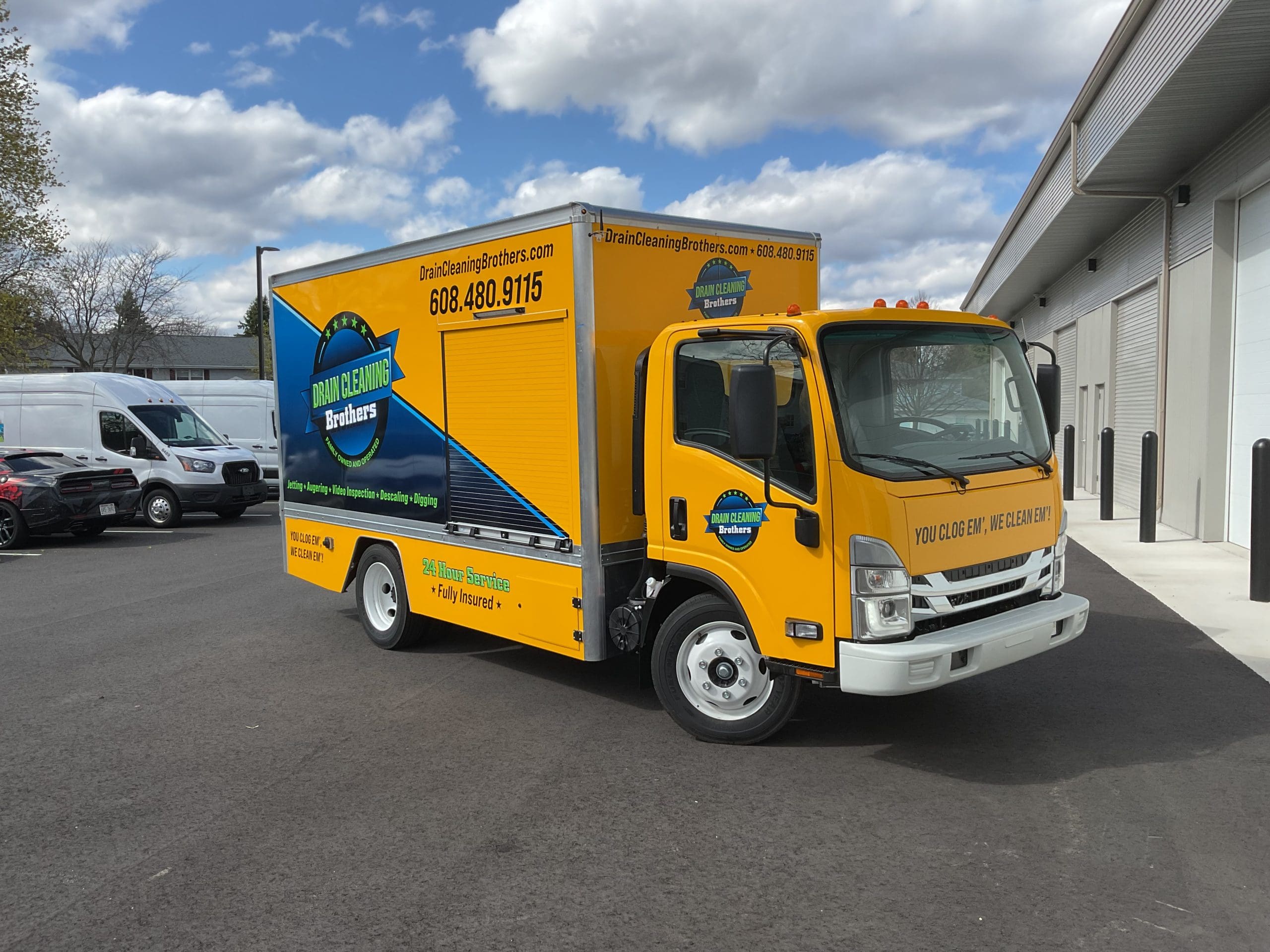 A yellow and blue Drain cleaning brothers hauling truck, promoting an plumbing business with fleet graphics, is parked outside a commercial building. The vehicle wrap features company details