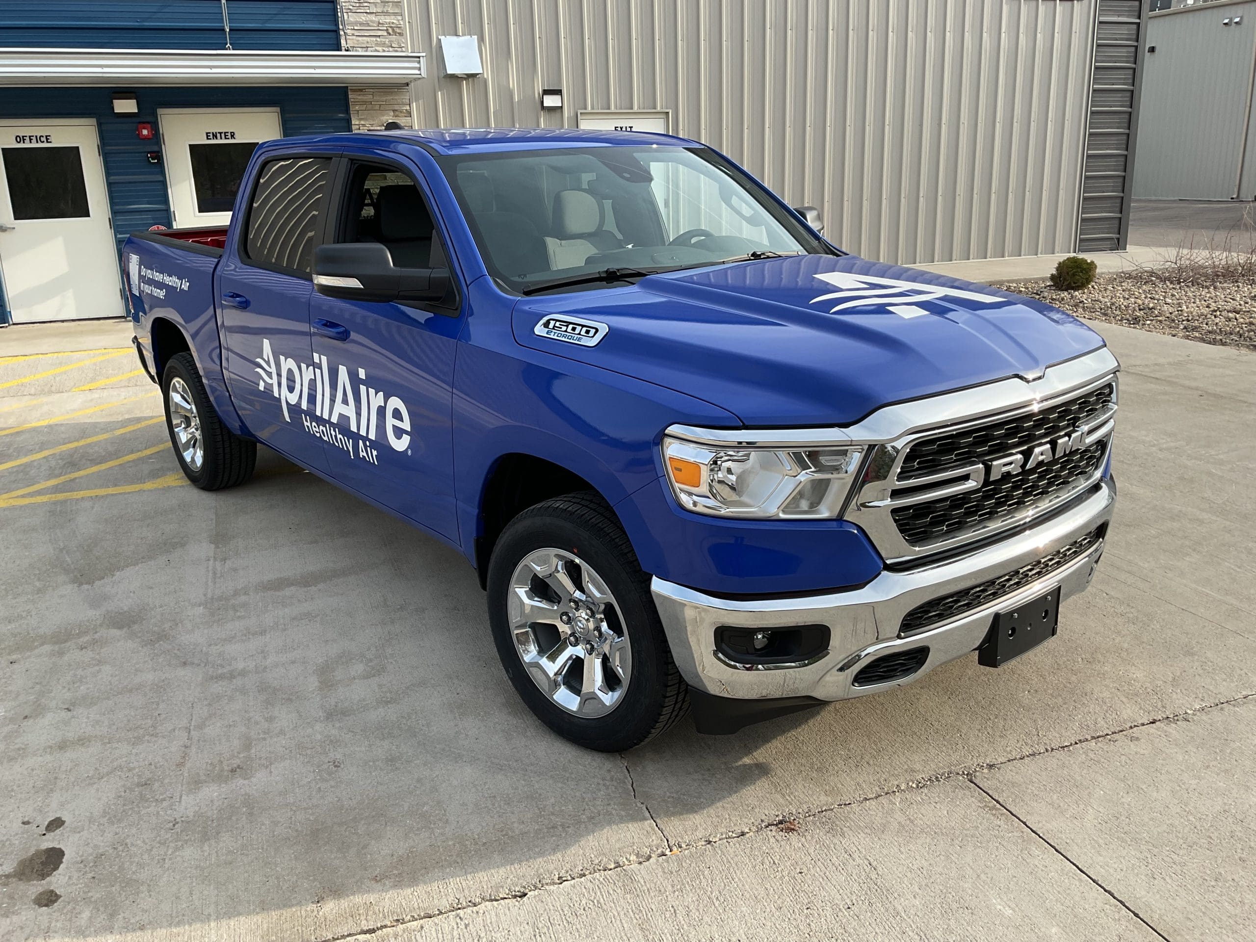 A dark blue April Aire pickup truck promoting a business with fleet graphics, is parked outside a commercial building.