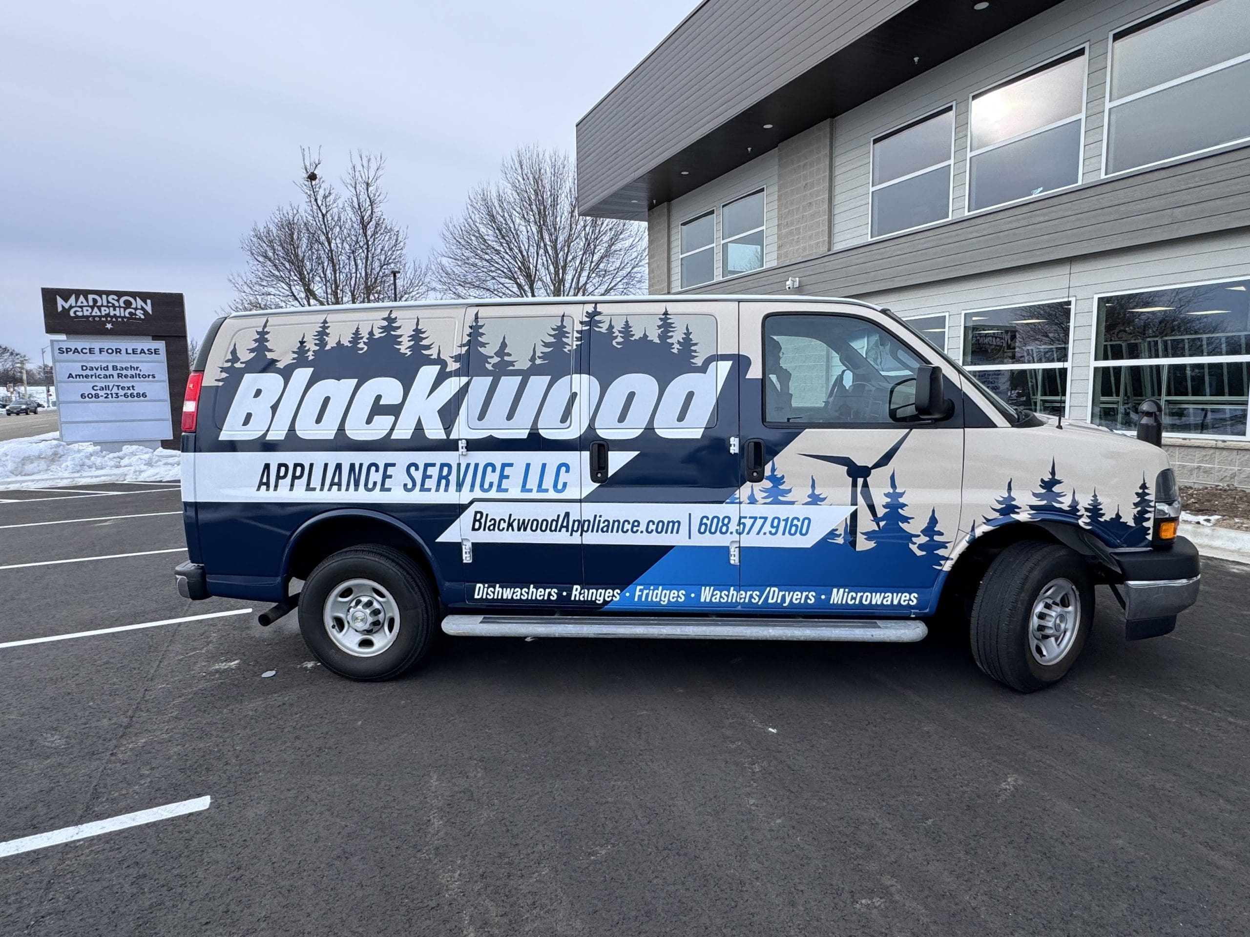 A white and blue Blackwood Appliance Service LLC van, promoting an appliance business with fleet graphics, is parked outside a commercial building. The van features company details and vibrant images of pines and mountains.