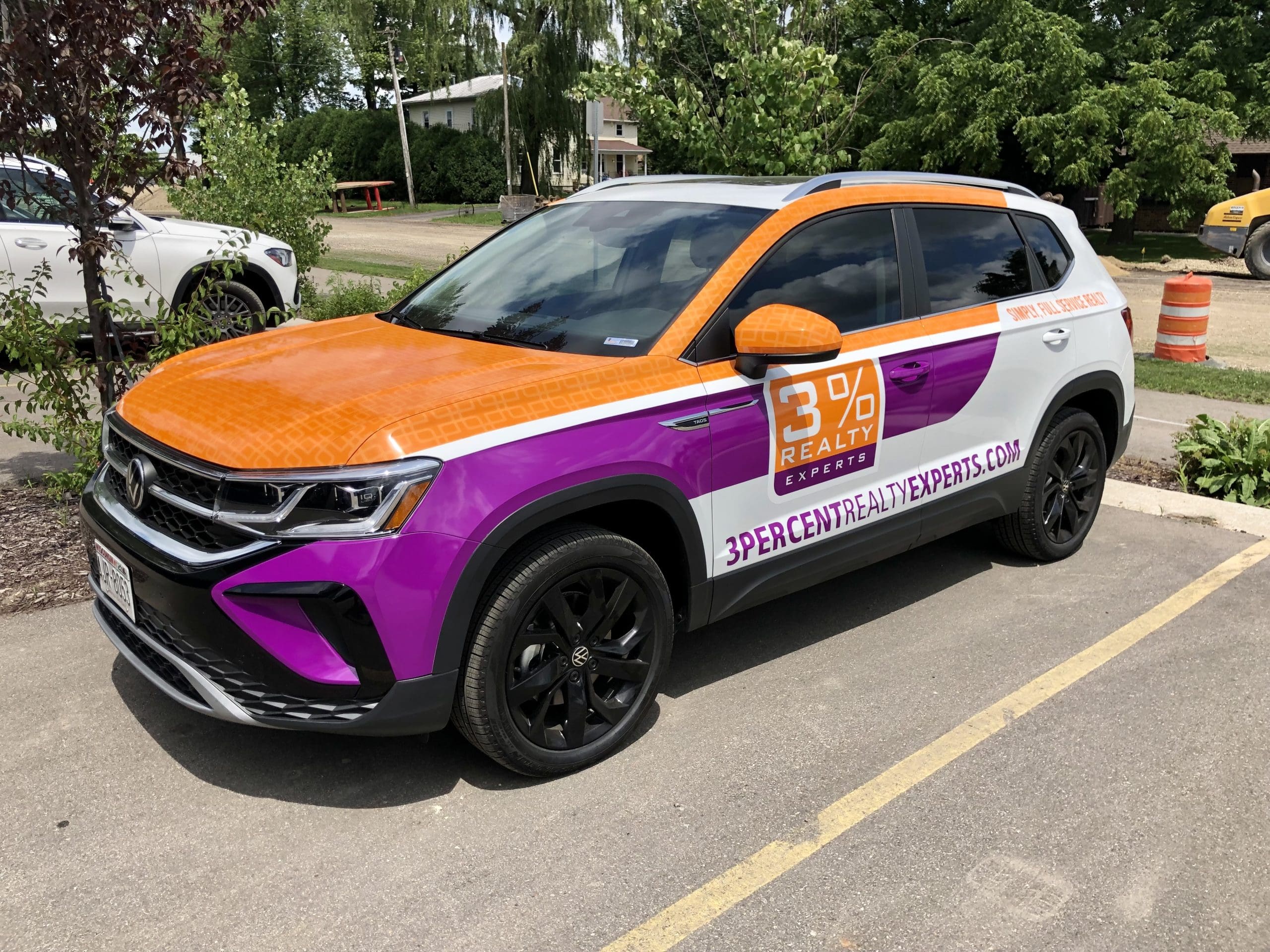 A white SUV with an orange hood and roof, and purple lower panels, is parked outside featuring 3% Realty Experts branding, website, and custom Fleet Wraps and Graphics. Trees and another car are visible in the background.