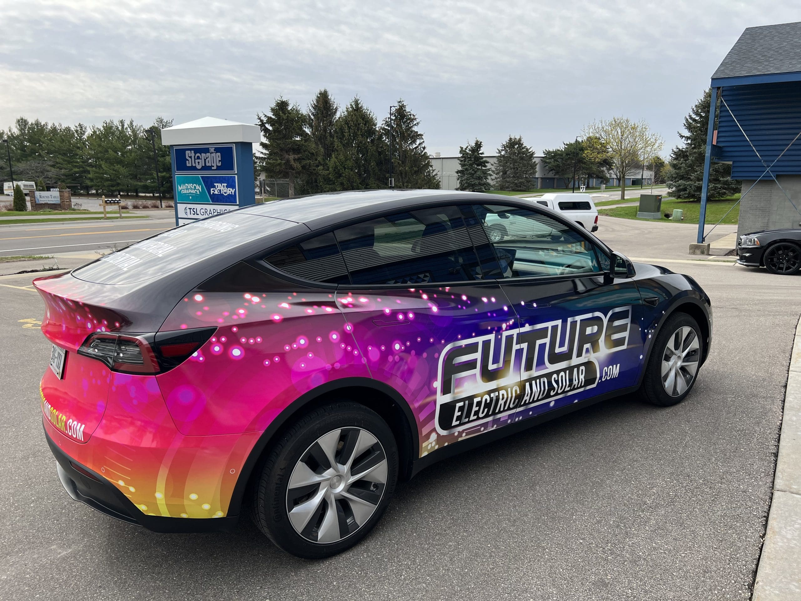 A car with a colorful gradient fleet wrap, displaying the text “FUTURE ELECTRIC AND SOLAR .COM,” is parked on a street near a blue building, with Fleet Wraps and Graphics making it stand out amid trees and signs in the background.