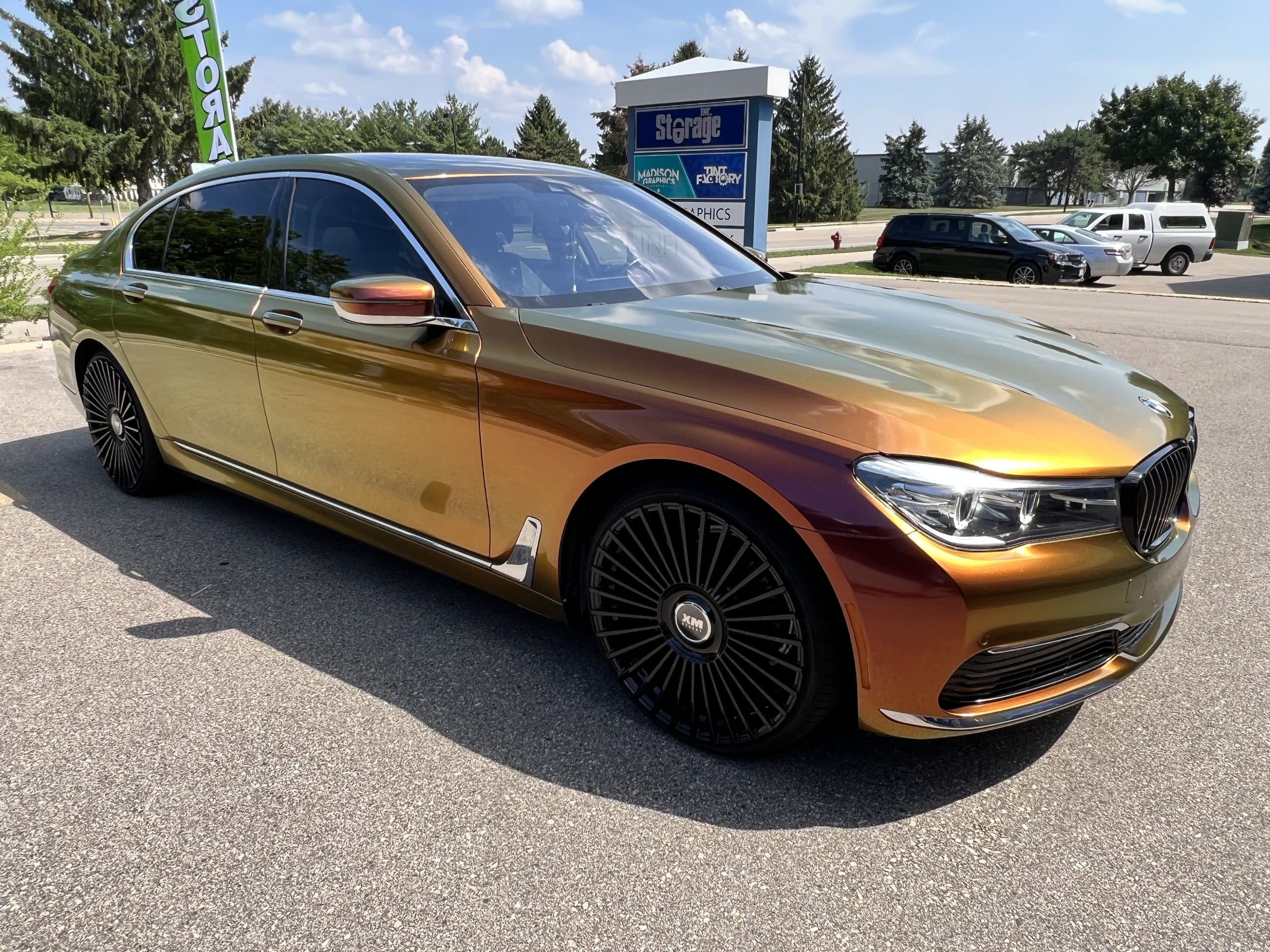 Sports car BMV with shiny gold with purple tint, sleek vehicle wrap designed my Madison Graphics. It is parked in a lot next to lots of trees