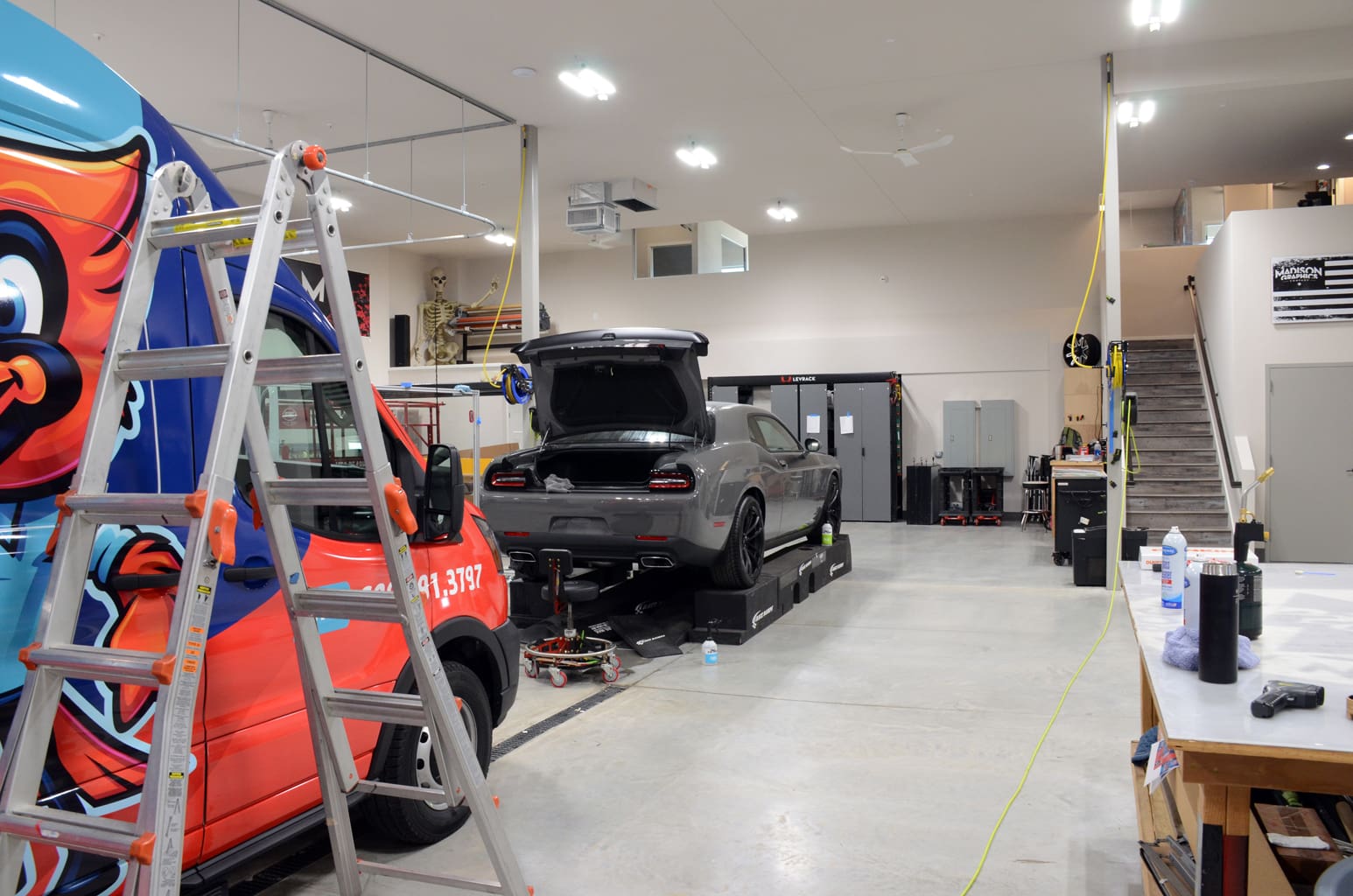 Design shop for fleet graphics with a colorful company van and a grey muscle car are being worked on you can see stairs to an upstairs section and a poster of Madison Graphics Company logo merged with an American flag