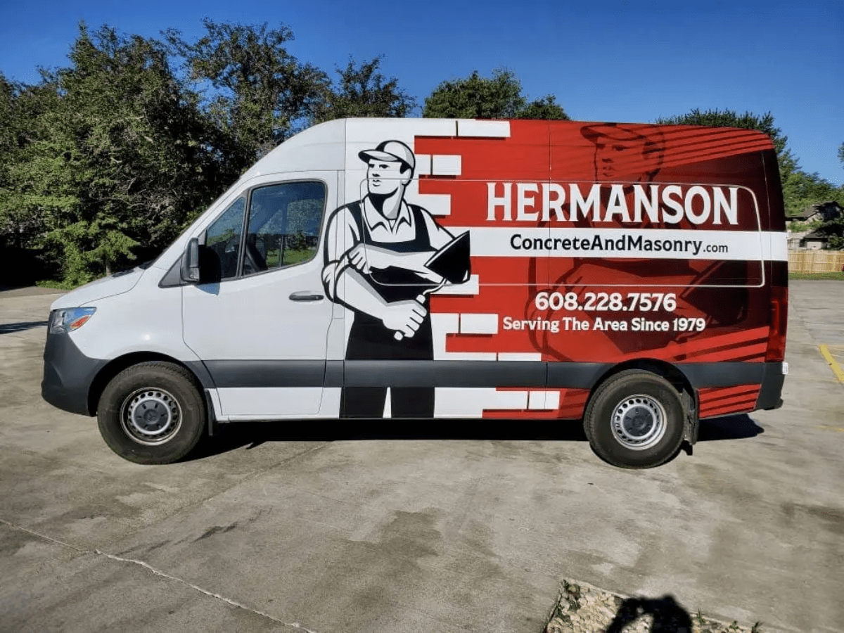 A Hermanson Concrete and Masonry van featuring fleet graphics and vehicle wraps, with a worker holding a trowel, contact info, and "Serving The Area Since 1979" on the side, is parked in a lot with trees in the background.