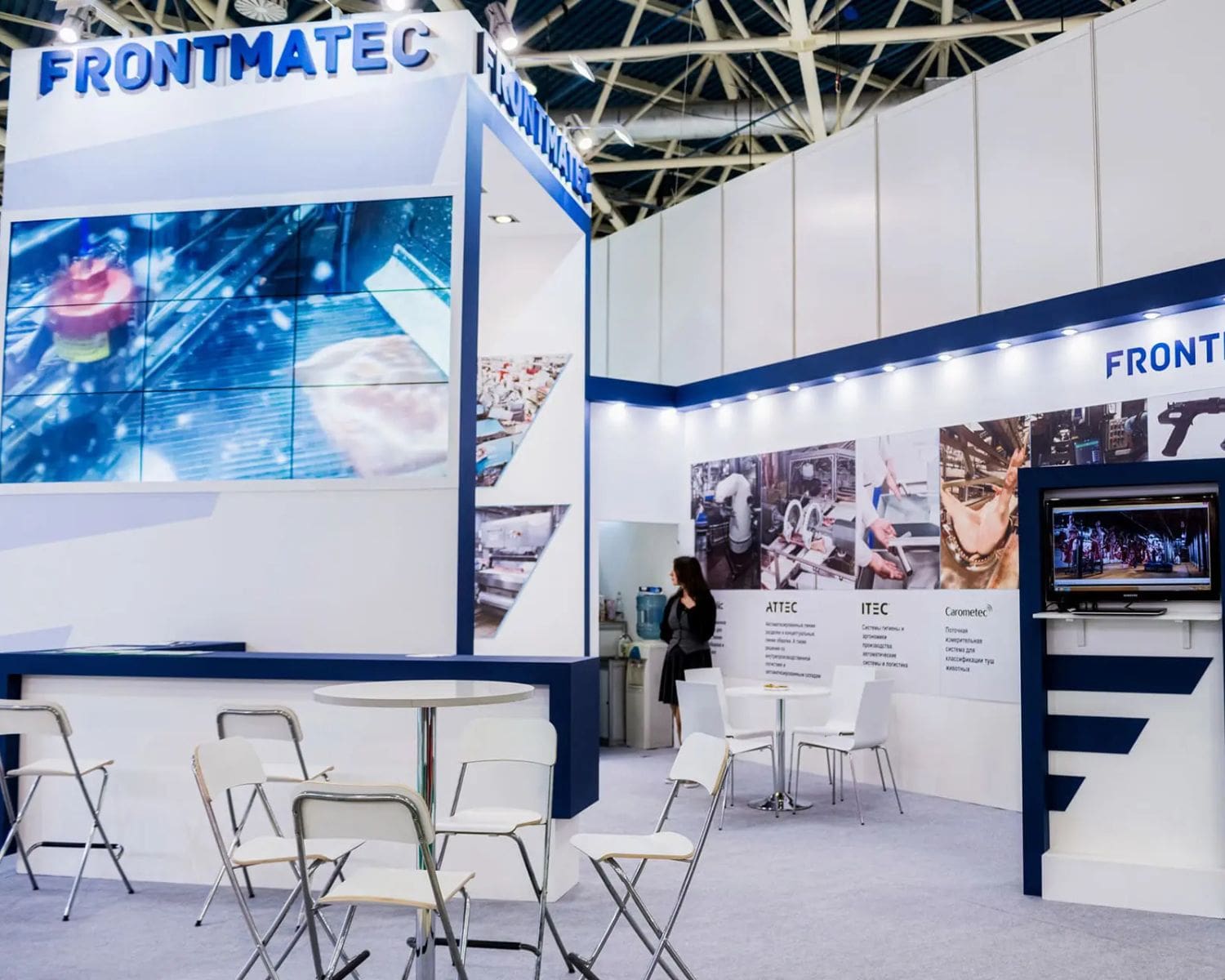 Trade show booth with FRONTMATEC branding, expert trade show graphics on display screens and posters, white tables and chairs. A person stands near the back wall featuring images and company info. Modern, well-lit, and professional setting.