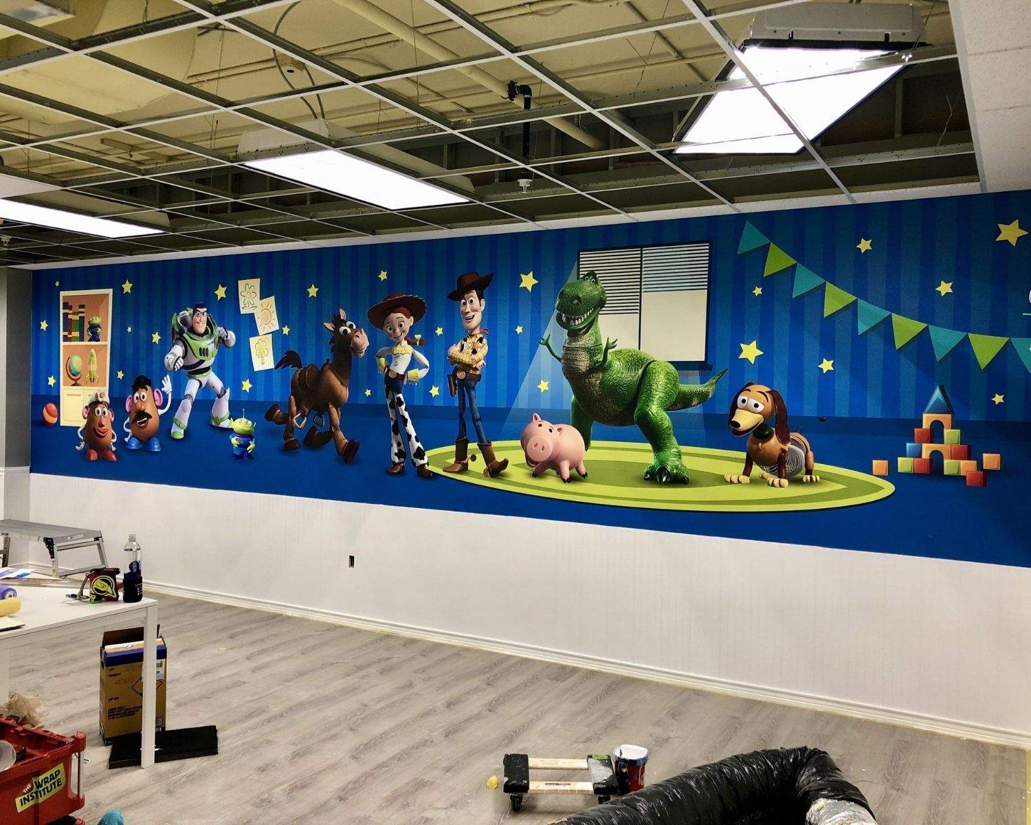 A colorful mural on a wall, inspired by Graphic Design Tips, depicts Toy Story characters—Buzz Lightyear, Woody, Jessie, Bullseye, Rex, Hamm, and Slinky Dog—on a blue background with stars and banners in a room under renovation.