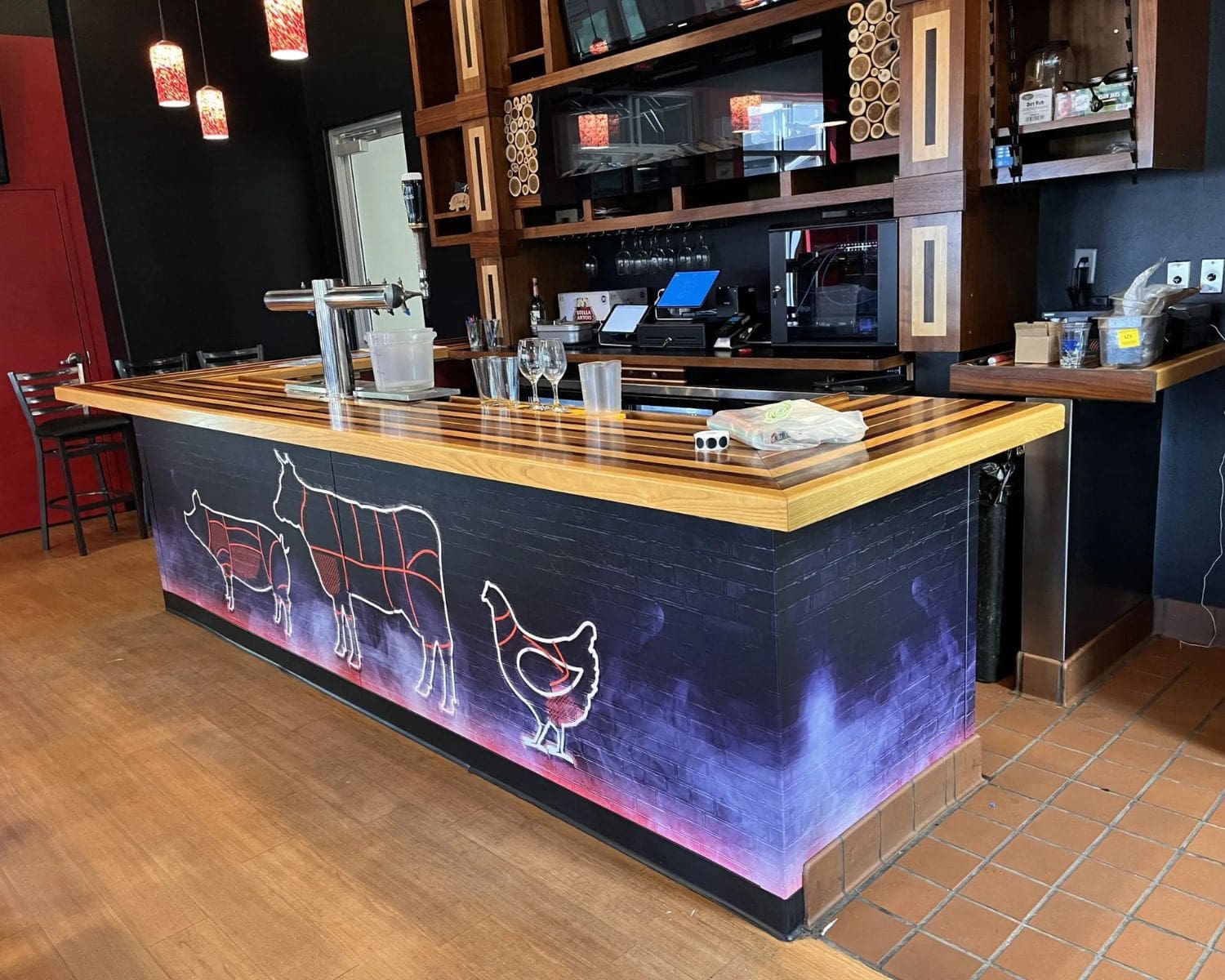 A restaurant bar counter with a wood top and animal outlines featuring butcher cuts, inspired by Graphic Design Trends. The wall behind displays shelves and drinks, while red pendant lights hang above for a modern touch.