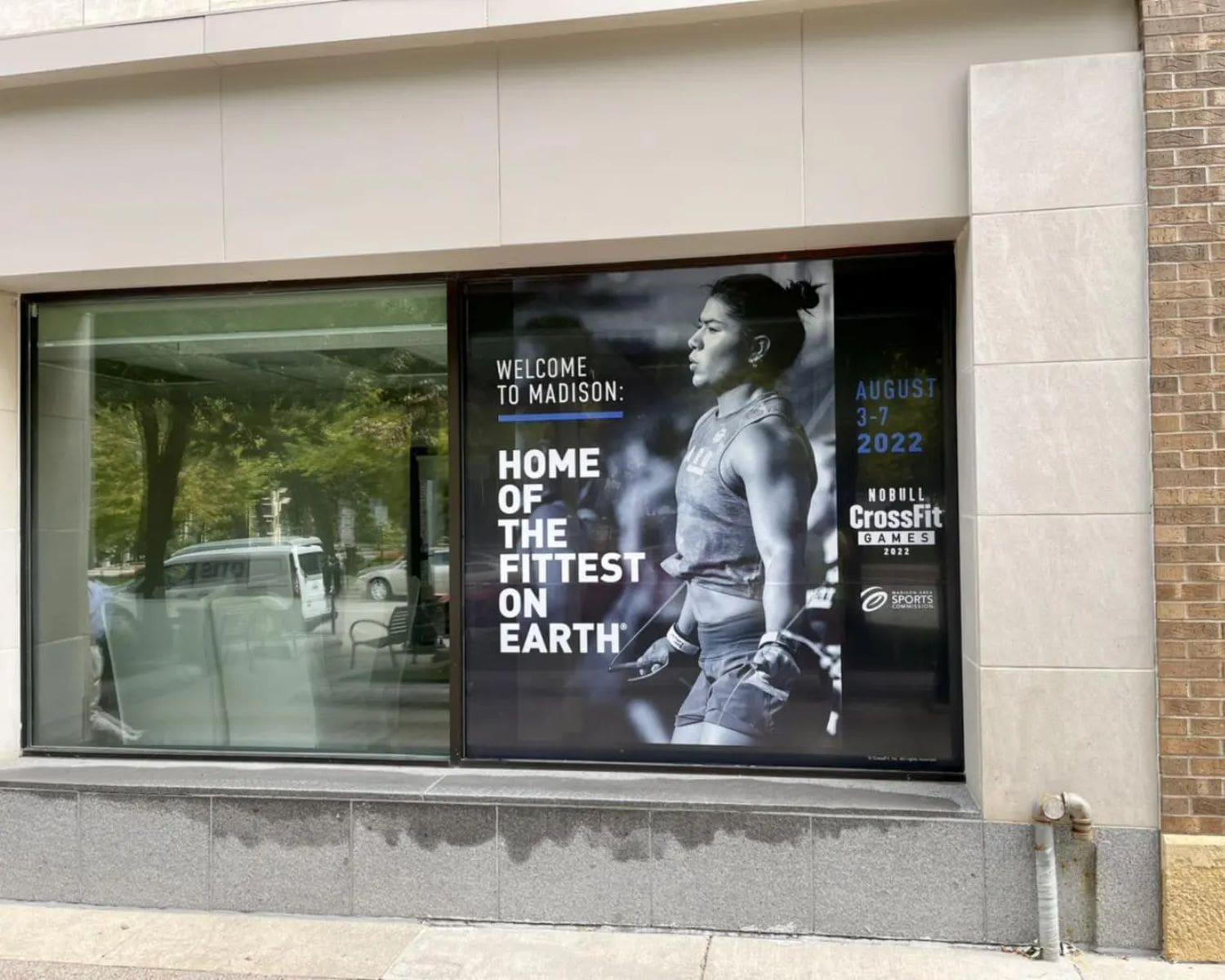 A large Window Graphics poster in a storefront window promotes the 2022 CrossFit Games in Madison, August 3-7, with the slogan Home of the Fittest on Earth and a black-and-white image of a focused female athlete.