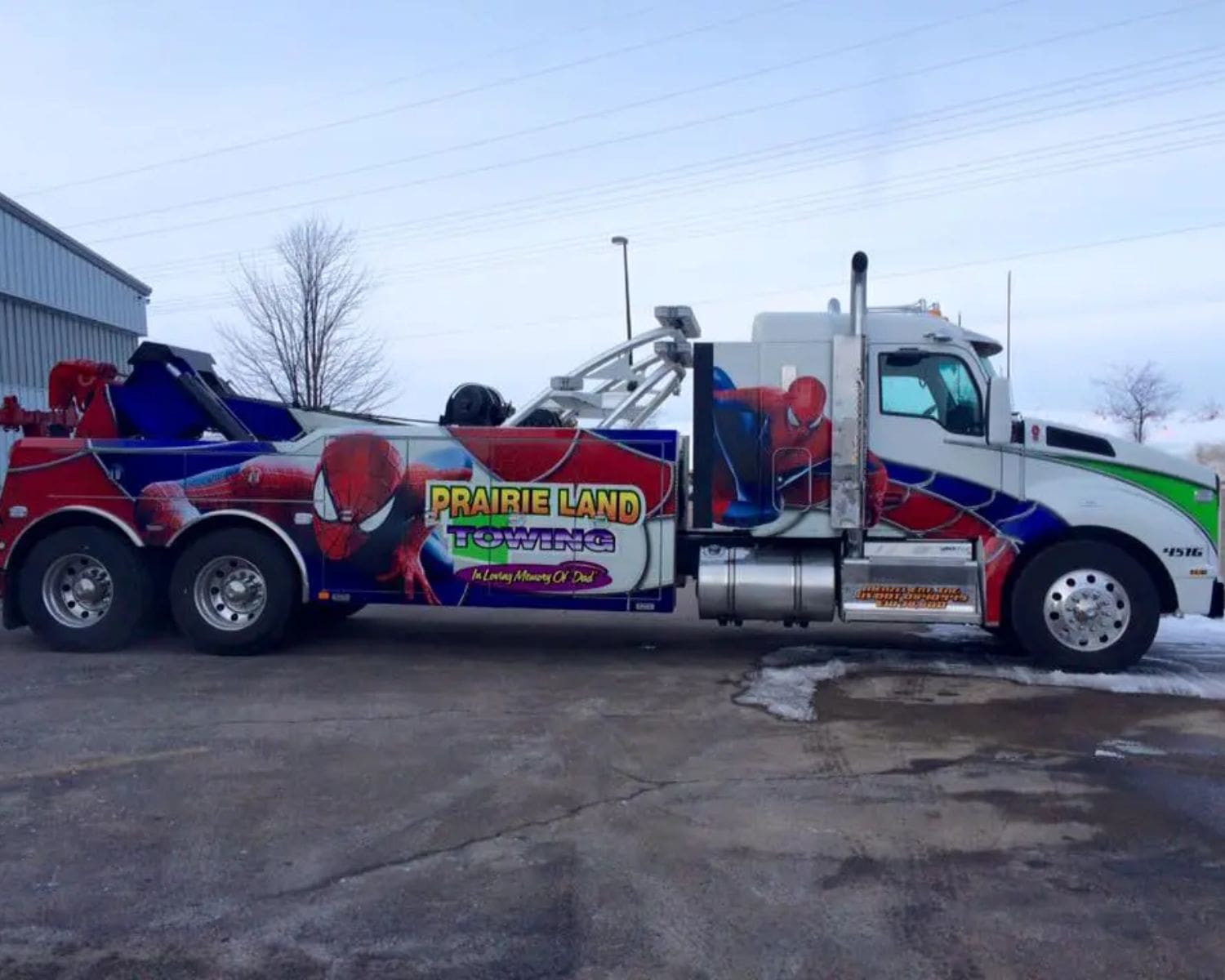 A large tow truck with colorful Spider-Man graphics and the words Prairie Land Towing on its side—showcasing Truck Fleet Graphics Madison—is parked on a snowy lot near a building.