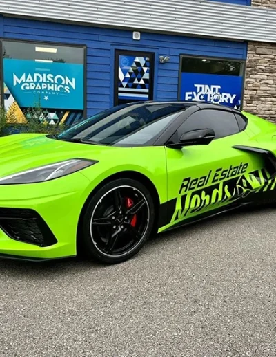 A bright green sports car featuring Fleet Wraps and Graphics, with bold black accents and Real Estate branding on the side, is parked outside a building displaying signage for MADISON GRAPHICS and TINT FACTORY.